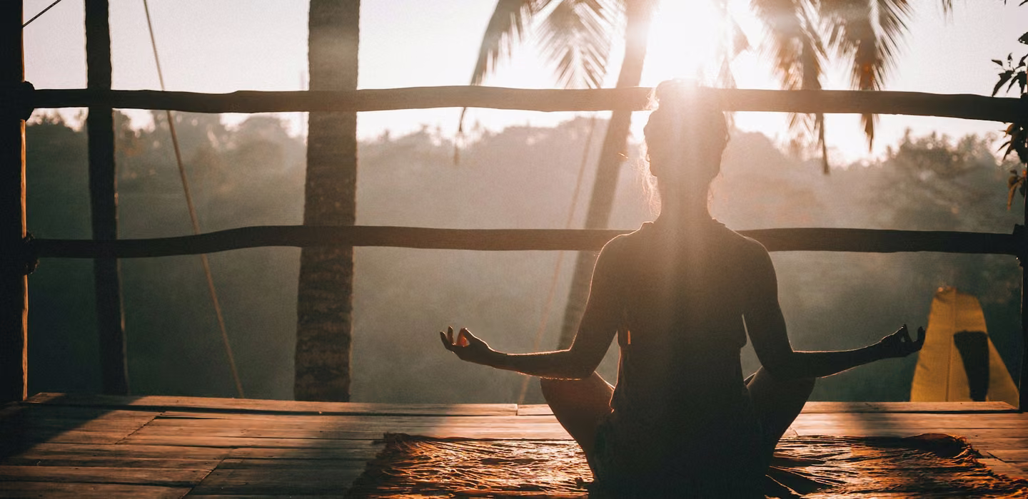 A guest meditating cross-legged on a wooden deck at sunrise, with palms behind